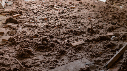 The floor shows a thick layer of dust and debris in a rundown building. Pieces of wood and other materials scatter among the dirt, showing signs of neglect and decay.