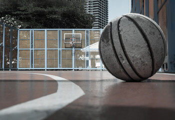An old basketball lies on the cement in the foreground of a half-outdoor basketball court that features a basketball hoop, fiberglass backboard, and a blue steel fence. Copy space.
