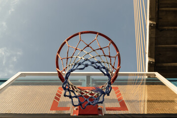 A low-angle view captures an orange metal basketball rim and net, mounted on a fiberglass basketball backboard, set against a clear blue sky. Space for text.