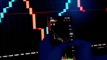Hands in blue gloves holding smartphone with stock market charts.