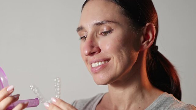 Woman uses dental trays for teeth whitening at home. Girl pours peroxide solution from syringe and applies on teeth, smiling to camera. Neutral background. Female holds trays in a purple case.