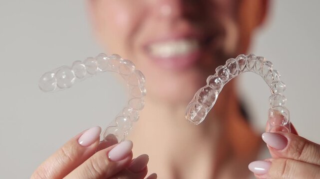 Woman uses dental trays for teeth whitening at home. Girl pours peroxide solution from syringe and applies on teeth, smiling to camera. Neutral background. Female holds trays in a purple case.