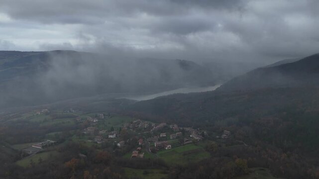 Slow drone pulling out from Mignano Dam, soaring through through low-hanging clouds in autumn mist in Arda Valley vibrant fall foliage of Apennine forests, toward hilltop hamlet of Castelletto, Italy
