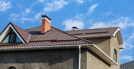 The roof of a house made of brown metal profile against a blue sky with white clouds