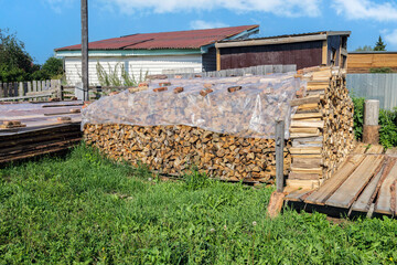A pile of firewood, covered with plastic, lies on a grassy field in a Siberian village. The scene is peaceful and serene, with the firewood and plastic creating a contrast with the green grass.
