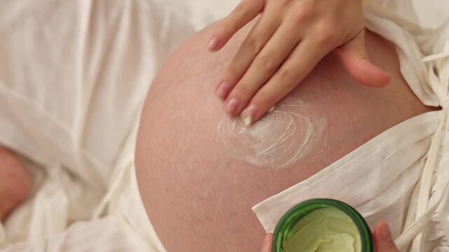 Pregnant woman lies on bed and applies cream on belly. Female in last month of pregnancy uses anti stretch mark cream from jar. She takes product with finger and gently massages abdomen. Body care rou