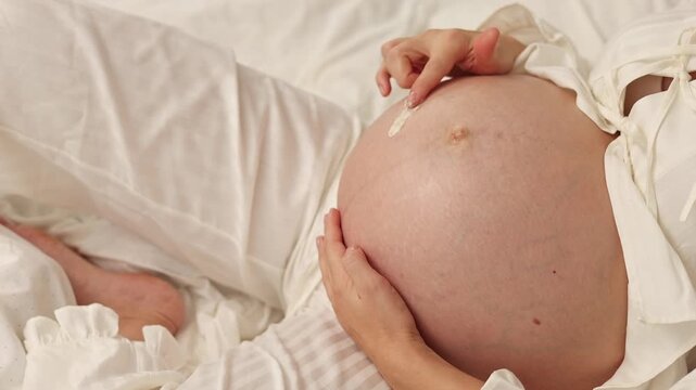 Pregnant woman lies on bed and applies cream on belly. Female in last month of pregnancy uses anti stretch mark cream from jar. She takes product with finger and gently massages abdomen. Body care rou