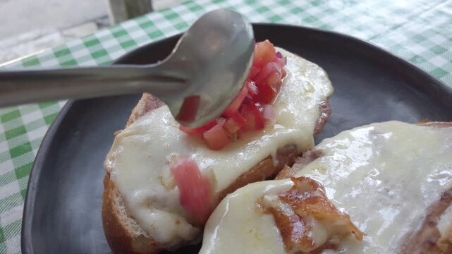A close-up of a spoon topping traditional Molletes (bread with beans and melted cheese) with fresh tomato and onion salsa.