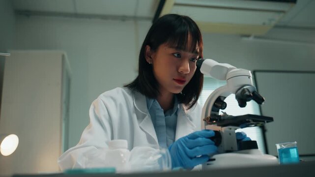 Young asian female microbiologist analyzing a sample through a microscope. She is wearing a lab coat and working on a scientific experiment