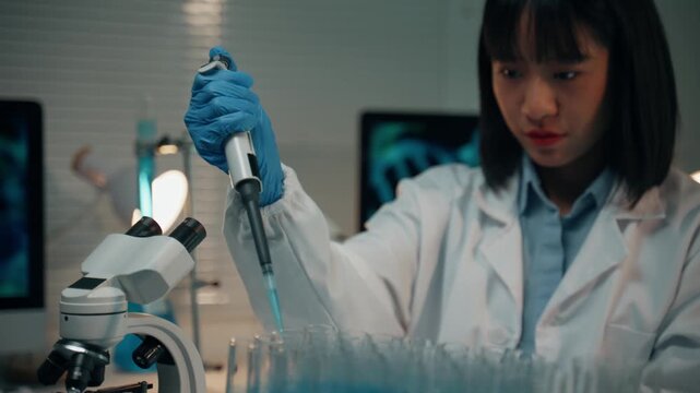 Focused young Asian research scientist using a micropipette to carefully transfer blue liquid into a row of test tubes during an experiment