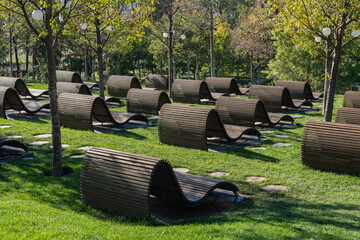 Modern, curved wooden benches arranged on green grass among young trees, creating unique, artistic and relaxing outdoor space. Recreation area is in Oblaka Park. Krasnodar City Park or Galitsky Park