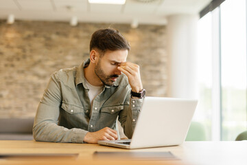 Young man sitting in an office feeling overwhelmed and stressed while using a laptop