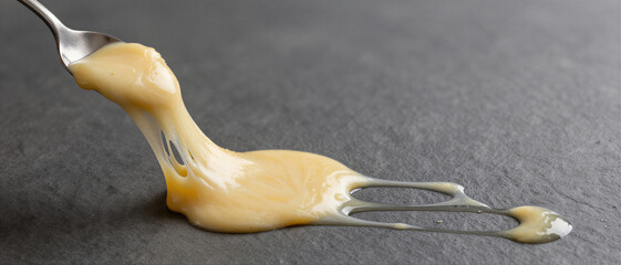 Thick melted cheese being stretched by a metal spoon on a dark slate background. Culinary food preparation concept.