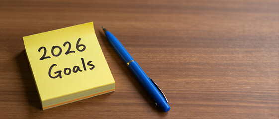 2026 Goals handwritten on a yellow sticky note next to a blue pen on a wooden desk. New Year's Resolution.