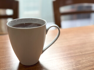 Obraz premium Steaming hot tea in a white ceramic cup on a wooden table with soft natural light and copy space. Cozy morning beverage scene in a café interior, minimal lifestyle concept.