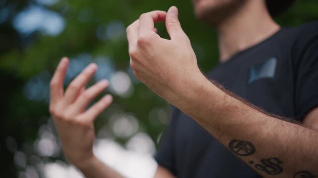 Tattooed Arms Caucasian Man Gesturing Emphatic, Expressive Finger Movements And Forearm Ink Visible Against Leafy Park Backdrop, Communicative Storytelling Posture And Dynamic Hand Language