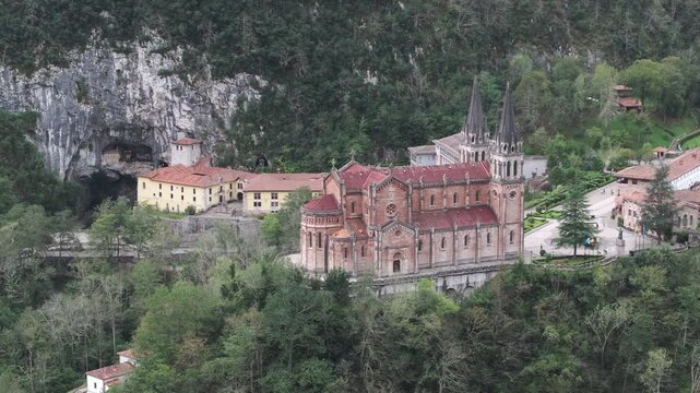 Aerial drone shot of the Basilica of Covadonga surrounded by dense forest and limestone cliffs, iconic religious landmark and pilgrimage site set deep in the mountains of Asturias, northern Spain