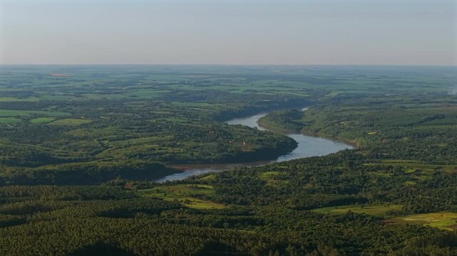 Cinematic high-altitude right tracking shot looking at the sinuous curves of the Paran&aacute; River border between Argentina and Paraguay, surrounded by vast jungle landscapes and hazy horizon.