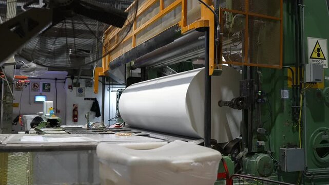 Cellulose paper jumbo roll mounted on an automated converting line inside a paper factory, showing the smooth white web, green machine frame, safety guards and surrounding control equipment.