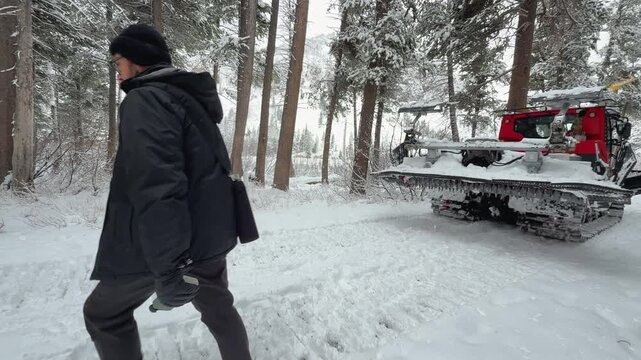 Man in a dark winter jacket walks past a red snowcat on a snowy forest road, showing heavy equipment maintaining mountain access in deep winter.