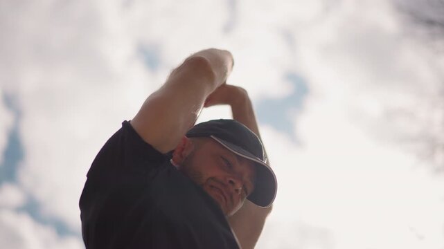 man adjusting cap under cloudy sky, lowangle backlit portrait showing strap over shoulder, focused expression as he stretches shoulder, tightens strap and scans horizon, sweat sheen on skin, casual