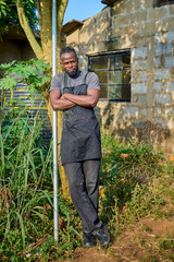 An African male farmer wearing an apron stands with folded arms beside a tree and metal pole in a rural farm environment near a simple building.