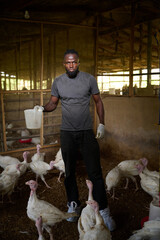 A Nigerian male farm worker stands inside an indoor poultry house holding a plastic container as white turkeys gather around him on the dirt floor near wooden enclosures.