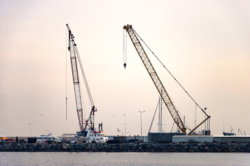 Fototapeta premium Harbor scene with red-white vessel crane and yellow dock crane—framed by rocky shoreline, flying seagulls, and maritime infrastructure in a coastal industrial setting under overcast daylight.