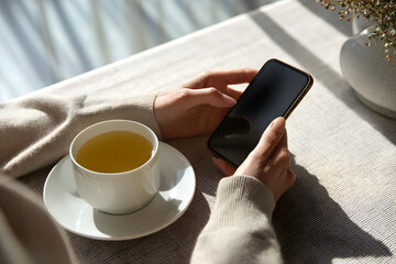 Person holds a smartphone and drinks tea at a table during daylight hours
