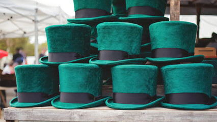 A stack of plush green St. Patrick’s hats sits on a wooden stall at an outdoor market in the morning. A gentle breeze causes the top hat to tilt slightly, showing shamrock patches