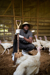 Young Nigerian male poultry farmer crouching inside an indoor turkey farm, holding a shovel while wearing gloves, a straw hat, and protective shoe covers as white turkeys surround him.