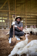 Young Nigerian male poultry farmer crouching inside an indoor turkey farm, holding a shovel while wearing gloves and protective shoe covers as white turkeys stand around him.