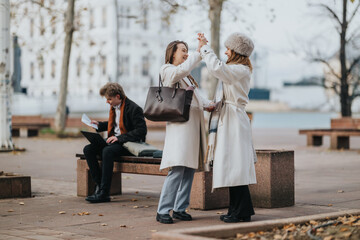 Two women in winter coats share a joyful high-five in a park on a chilly day. Other people sit and walk in the background, creating a casual urban scene.