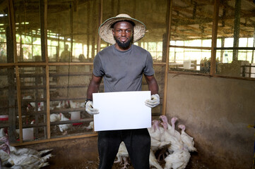 Young African male poultry farmer standing inside an indoor turkey farm, holding a blank white sign with gloved hands while wearing a straw hat and casual work clothes, with turkeys in the background.
