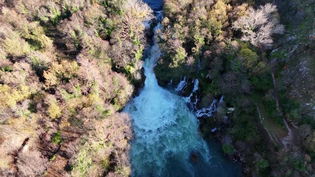 A drone lifts over Bilusića Buk waterfall, the Krka River, revealing the fan of cascading water. The camera continues over a dense forest and canyon, revealing the distant peaks of Dinara Mountain.
