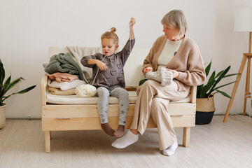 In a bright living room, a young girl plays with yarn while her grandmother knits. Both share a joyful moment on a simple wooden bench surrounded by plants