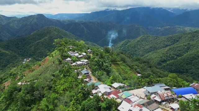 Push in overhead aerial gliding over rural houses toward rising smoke, revealing lush mountain ridges and deep valleys in Banaue, Ifugao, Philippines.