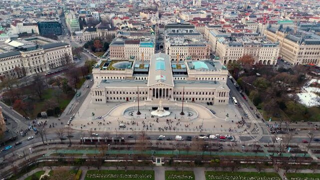 Aerial landscape drone shot panning over the Austrian Parliament Building, showing surrounding roads, moving traffic, and Vienna city blocks beneath clear winter sunlight and crisp seasonal shadows.