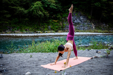 Asian woman practicing advanced yoga balance pose outdoors by a river, strength and mental wellness lifestyle in nature