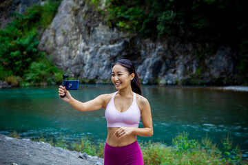 Fototapeta premium Smiling fitness influencer recording selfie video with smartphone gimbal outdoors by river