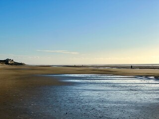 Quend-Plage, Authie Bay, France wide sandy beach at low tide with tidal pools and sunlit wet sand reflections, calm horizon under clear blue sky, minimalist seascape