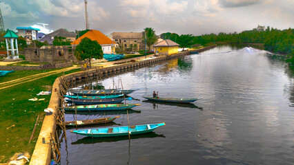 Aerial view of boats with vibrant colors resting by the waterfront near buildings with orange roofs under a cloudy sky, Abonnema, Port Harcourt, Nigeria.