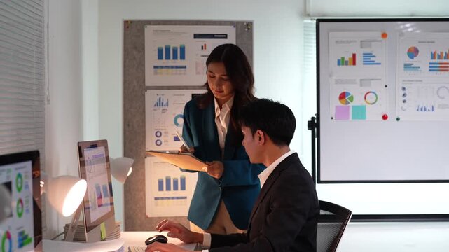 Two dedicated asian business colleagues working overtime, analyzing charts and graphs on a computer screen in a modern office, collaborating on a project to meet a deadline