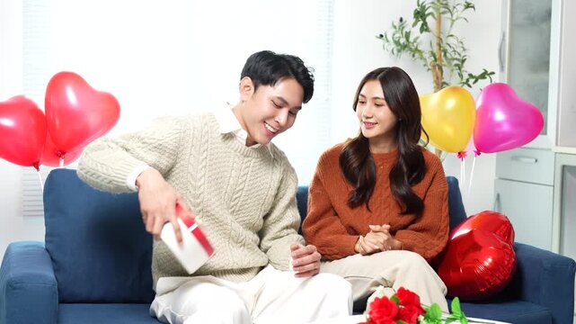 Happy young asian man giving his girlfriend a romantic surprise with a heart-shaped gift box while sitting on the couch in a living room decorated with red heart balloons for valentine's day