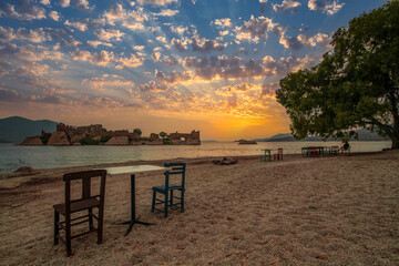 The diverse landscapes of Lake Bafa in T&uuml;rkiye's Aegean region include colorful sunsets, an island, a monastery and rock formations, and a lakeside relaxation area with tables and chairs.