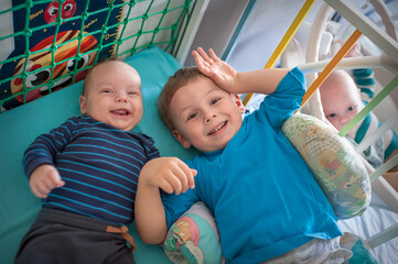 Two smiling brothers of different ages are lying in a crib together. One is smiling, and the other is waving. A third brother peeks from behind the crib bars.