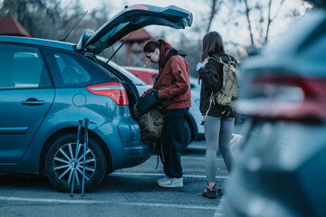 Two women bustle at a car trunk, packing backpacks for a hiking trip at a campsite. The scene shows travel gear and tents in the background, conveying outdoor adventure and preparation.