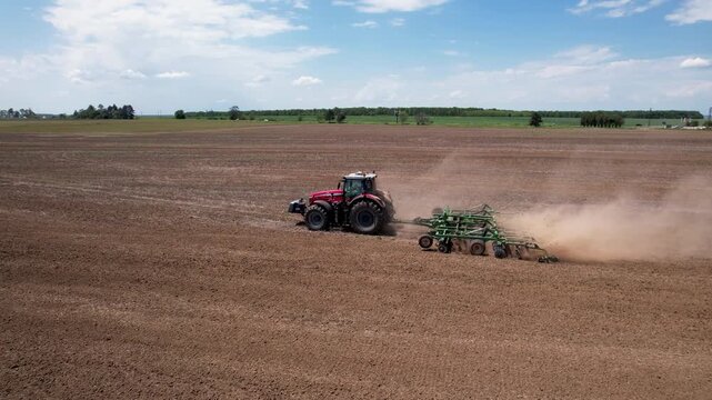 Aerial view of a red tractor pulling a green tiller, stirring up dust in a vast brown field under a partly cloudy sky, Silistra, Bulgaria.