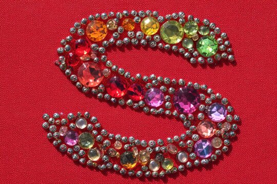 Close-up of letter "S" constructed from colorful jewels and surrounded by silver beads on red fabric - Powered by Adobe