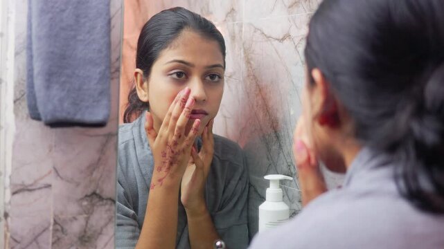 Indian woman in bathrobe, gently touching her cheeks and examining facial skin condition in front of a bathroom mirror.